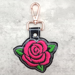 Puede incluir: Un llavero de cuero negro con una rosa rosa bordada sobre tela negra y hojas verdes. El llavero tiene un cierre giratorio de oro rosa.