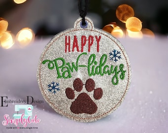 May include: Glittery round ornament with the words "HAPPY Paw-lidays" in red and green, a brown paw print, and blue snowflakes. The ornament has a black ribbon for hanging. The background is blurred with bokeh lights.