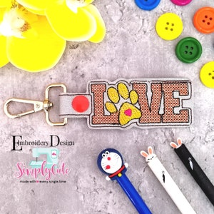 May include: A brown and white embroidered key ring with the word "LOVE" and a paw print in the centre. The paw print has a small red heart in the centre. The key ring has a gold metal clasp.