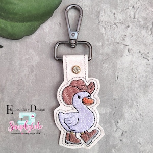 May include: A white leather keychain with a silver metal clasp. The keychain features an embroidered design of a purple duck wearing a brown cowboy hat and brown boots.