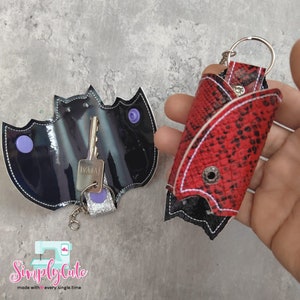 May include: Two keychains, one shaped like a bat and the other a snake skin design. Both are black with purple accents and have a silver key ring.