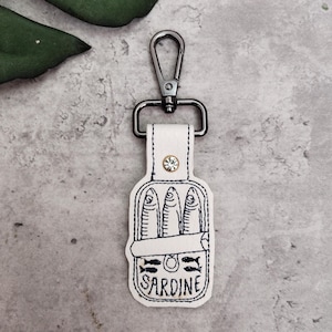 Könnte beinhalten: Weißer Leder-Schlüsselanhänger mit Sardinenbüchsen-Motiv und schwarzen Stickereidetails. Der Schlüsselanhänger hat einen Metallverschluss und einen kleinen Edelstein. Das Sardinenbüchsen-Design zeigt drei Fische und das Wort "Sardine".