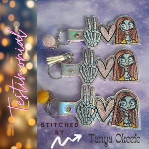 May include: Three pink and white keychains with a peace sign, heart, and a cartoon character with long red hair. The keychains are embroidered with white thread and have a silver metal ring. The text "STITCHED BY Tanya Okeefe" is visible at the bottom of the image.