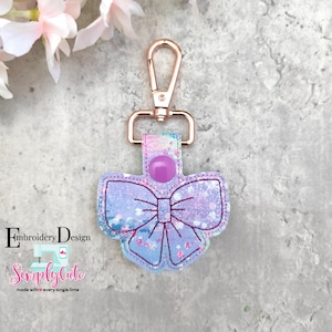 May include: A purple and blue glitter bow keychain with a rose gold swivel clip. The bow has a purple button in the center.