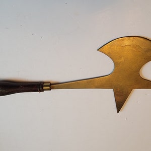 Puede incluir: Una herramienta antigua de latón con un mango de madera oscura y una hoja única en forma de estrella. El mango mide aproximadamente 13 cm de largo, y la hoja de latón tiene un diseño asimétrico distintivo. La herramienta parece bien conservada y muestra signos de edad.