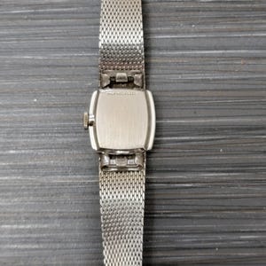May include: A silver watch with a square face and a silver mesh band. The back of the watch has the text "14K GOLD FILLED" engraved on it.