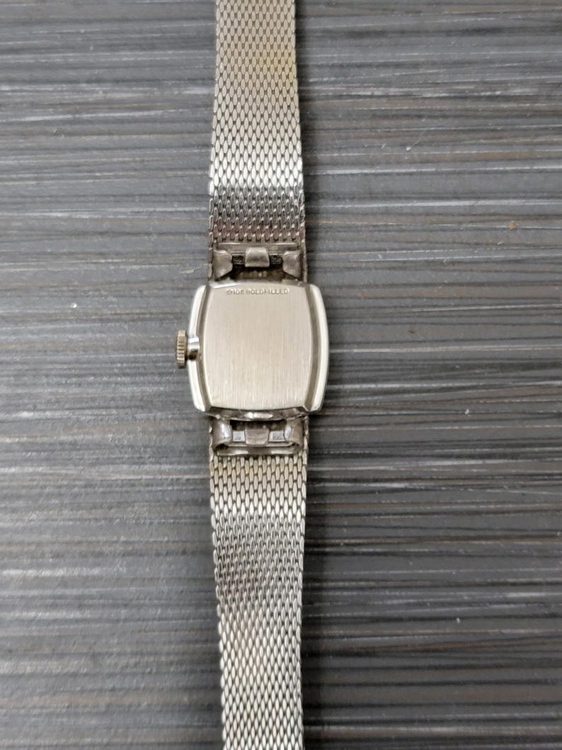 May include: A silver watch with a square face and a silver mesh band. The back of the watch has the text "14K GOLD FILLED" engraved on it.