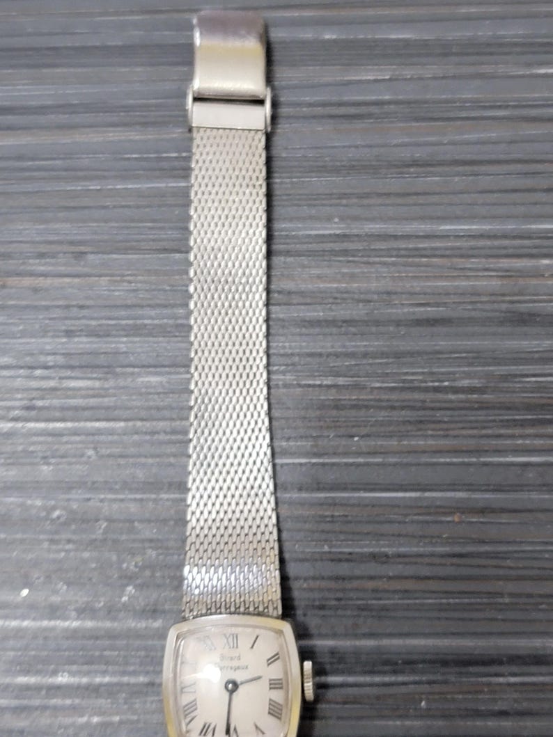 May include: A silver watch with a square face and a silver mesh band. The watch face has Roman numeral hour markers and a white background. The watch is a vintage style.