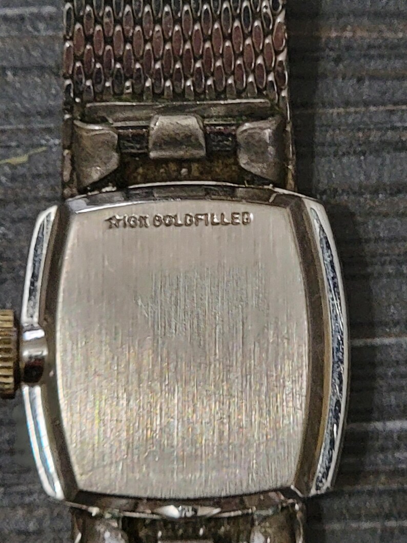 May include: A silver watch with a square face and a metal band. The back of the watch has the words "10K Gold Filled" engraved on it.