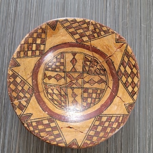 May include: A round, antique ceramic bowl with a geometric design in shades of brown and red. The bowl features a central circular pattern and a cracked surface, suggesting age and use. The bowl's design includes diamond and square patterns.