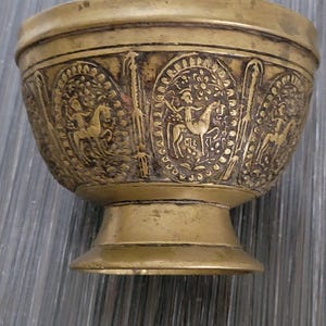 May include: A small, gold-coloured brass bowl with an intricate design. The bowl has a wide, round base and a flared lip. The design features a repeating pattern of horses and riders.