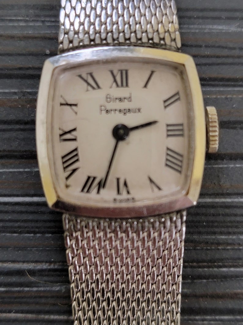 May include: A silver watch with a square face and a silver mesh band. The face has Roman numeral hour markers and the text "Girard Perregaux" in black.