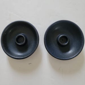 May include: Two circular, matte black candle holders with a wide, shallow, bowl-like base and a central cylindrical opening designed to hold a single taper candle. The surface of the holders appears textured and uniform in colour.