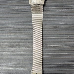 May include: A silver watch with a square face and a silver mesh band. The watch face has Roman numerals and the text "Girard Perregaux" on it.