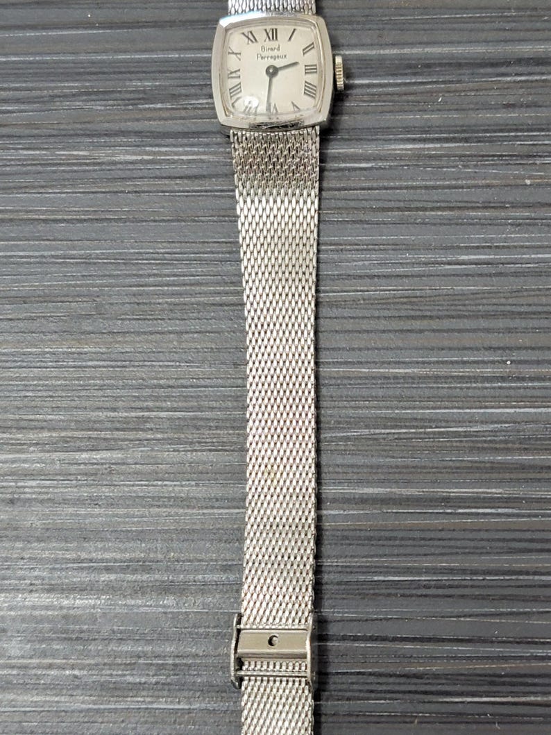 May include: A silver watch with a square face and a silver mesh band. The watch face has Roman numerals and the text "Girard Perregaux" on it.