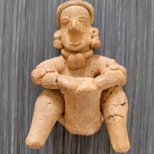 Pode incluir: Pequena figura de cerâmica de cor castanha clara de uma figura sentada com os braços cruzados. A figura tem uma cabeça arredondada, um nariz proeminente e um design simples e estilizado. A figura parece ser feita à mão e tem uma textura ligeiramente áspera.