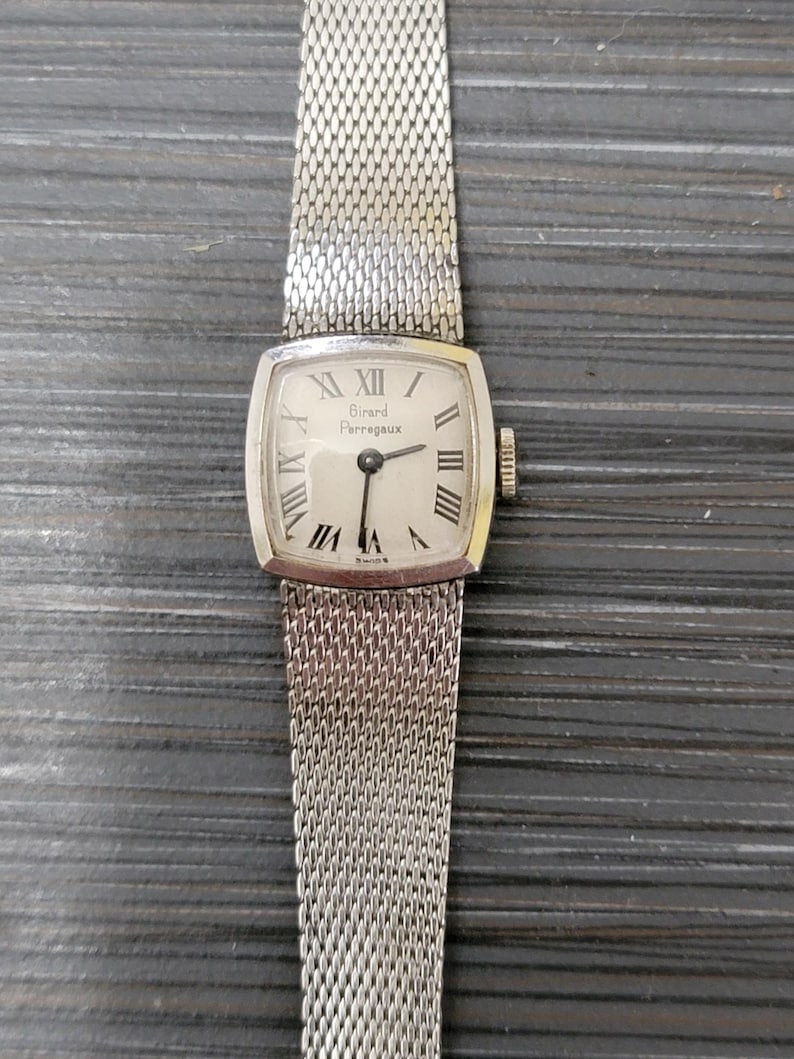 May include: A silver wristwatch with a square face and a silver mesh band. The watch face has Roman numeral hour markers and the text "Girard Perregaux" printed on it.