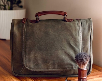Puede incluir: Una bolsa de viaje de lona verde con asa de cuero marrón y cierres de cremallera. Una navaja de afeitar de seguridad plateada y una brocha de afeitar marrón están en primer plano.