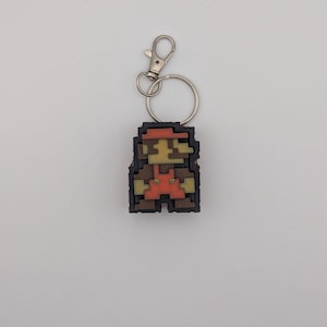 Könnte beinhalten: Ein pixeliger Schlüsselanhänger mit einer Figur in roter Mütze und Latzhose. Der Schlüsselanhänger hat einen silbernen Verschluss und Ring. Die Figur ist im klassischen Videospielstil gehalten, mit braunen, roten und beigen Farben.