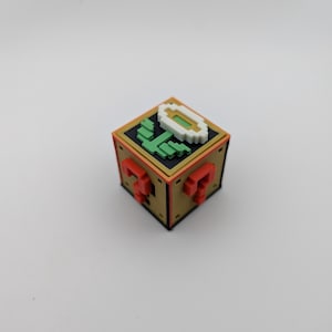 Könnte beinhalten: Ein pixeliger Würfel mit goldenem, rotem und schwarzem Design. Die Oberseite zeigt eine grüne Pflanze und eine weiße Blume. Die Seiten haben rote Fragezeichen. Dieser Artikel ist wahrscheinlich ein Sammler- oder Dekorationsartikel.