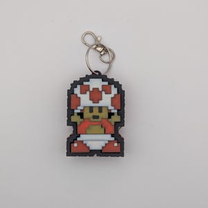 Könnte beinhalten: Ein pixeliger Schlüsselanhänger mit einer Figur aus der Super Mario Bros.-Reihe. Der Schlüsselanhänger hat eine schwarze Umrandung, eine weiß-rote Pilzkappe und eine rote Weste. Er ist an einem silbernen Schlüsselring befestigt.