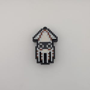 Könnte beinhalten: Ein pixeliger Tintenfisch-Anhänger in Schwarz, Weiß und Braun. Der Anhänger hat oben eine kleine Schlaufe zur Befestigung. Das Design erinnert an Retro-Videospielgrafiken. Der Körper des Tintenfischs ist weiß mit braunen und schwarzen Akzenten.