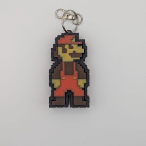 Könnte beinhalten: Ein pixeliger Schlüsselanhänger mit der Figur Mario aus dem Videospiel. Der Anhänger hat eine schwarze Umrandung, einen roten Hut und Latzhose, ein braunes Hemd und beige Haut. Er ist an einem silbernen Metallring befestigt.