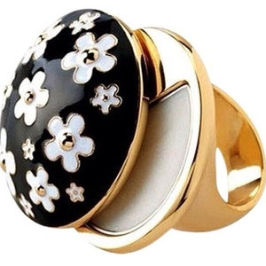 Op de afbeelding: Een goudkleurige ring met een zwarte ovale bovenkant versierd met witte madeliefachtige bloemen. De bovenkant van de ring kan worden geopend om een wit interieur te onthullen. De ring heeft een brede band en een gepolijste afwerking.
