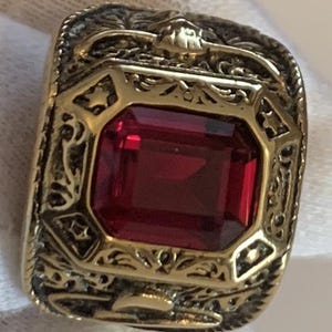 Può includere: Un anello dorato con una grande gemma rosso scuro rettangolare al centro. L'anello presenta un design ornato e dettagliato attorno alla gemma e sulla fascia, con motivi floreali e a stella. L'anello è su sfondo bianco.