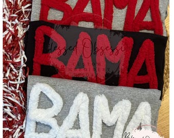 May include: Three folded t-shirts stacked vertically. The top shirt is heather gray with red fuzzy chenille letters spelling BAMA. The middle shirt is black with red fuzzy chenille letters spelling BAMA. The bottom shirt is heather gray with white fuzzy chenille letters spelling BAMA.