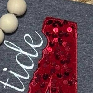 May include: A dark gray fabric surface featuring a red sequined letter A patch and white embroidered cursive text that reads tide. A string of light-colored wooden beads is positioned along the upper left edge of the gray fabric material.