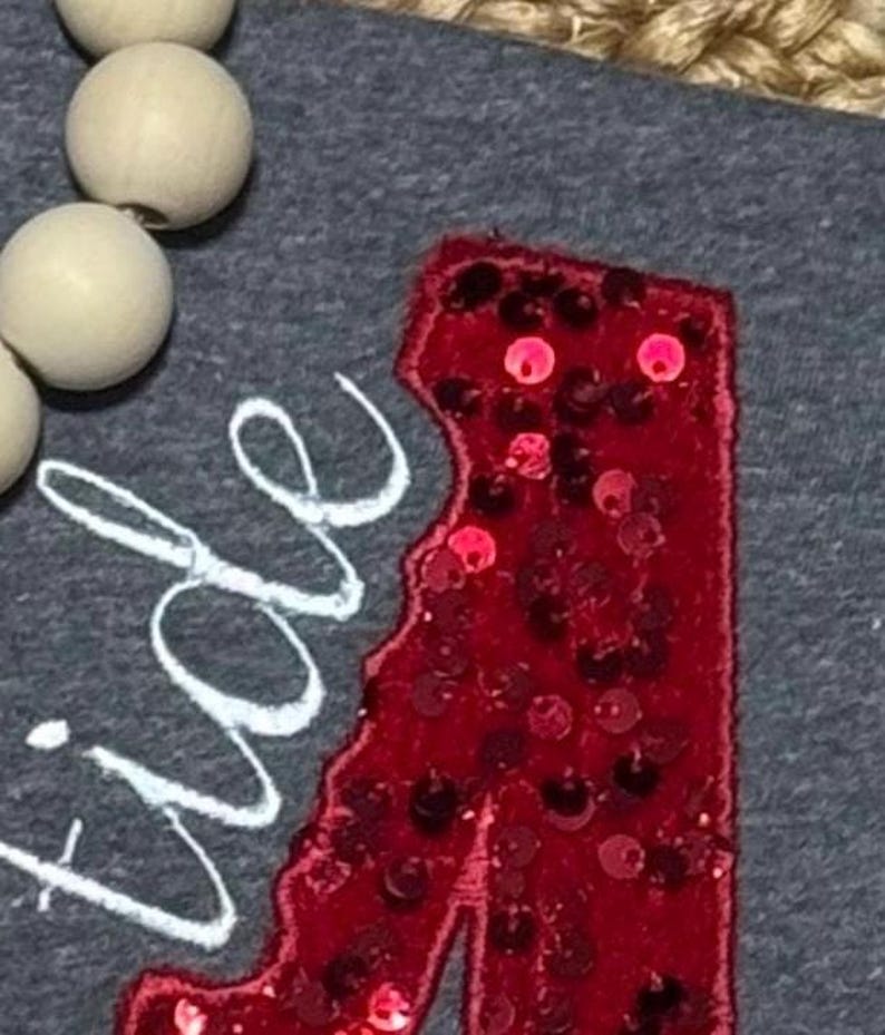 May include: A dark gray fabric surface featuring a red sequined letter A patch and white embroidered cursive text that reads tide. A string of light-colored wooden beads is positioned along the upper left edge of the gray fabric material.