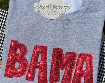 May include: A heather gray crewneck sweatshirt featuring the word BAMA in large, red, sequined block letters across the chest. A circular white tag is attached to the neckline with the text Blessed Obsessions Custom Embroidery & More.