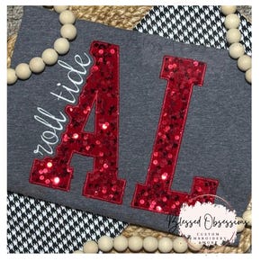 May include: A dark gray t-shirt featuring large, red sequined letters spelling AL, with the white embroidered text roll tide positioned to the left. The design is accented by a border of light-colored wooden beads and a black and white houndstooth pattern.