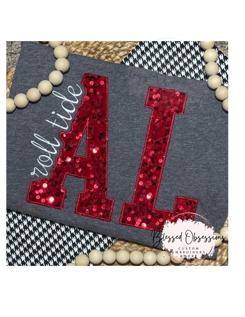 May include: A dark gray t-shirt featuring large, red sequined letters spelling AL, with the white embroidered text roll tide positioned to the left. The design is accented by a border of light-colored wooden beads and a black and white houndstooth pattern.