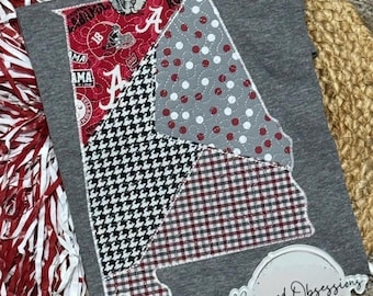 May include: A heather gray t-shirt featuring a large Alabama state-shaped applique. The design is divided into four sections with red patterned fabric, gray fabric with white dots, black and white houndstooth, and a red and black gingham check.