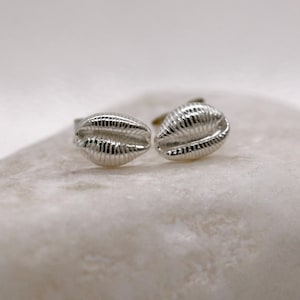 May include: A pair of silver cufflinks, each shaped like a seashell. The cufflinks have a textured surface with detailed ridges. They are displayed on a light-coloured, textured surface.
