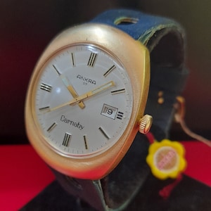 Könnte beinhalten: Eine goldfarbene Armbanduhr mit weißem Zifferblatt und schwarzen Zeigern. Die Uhr hat ein schwarzes Lederarmband und ein Datumsfenster bei 3 Uhr. Das Zifferblatt der Uhr zeigt "Ankra 07" und "Carnaby".