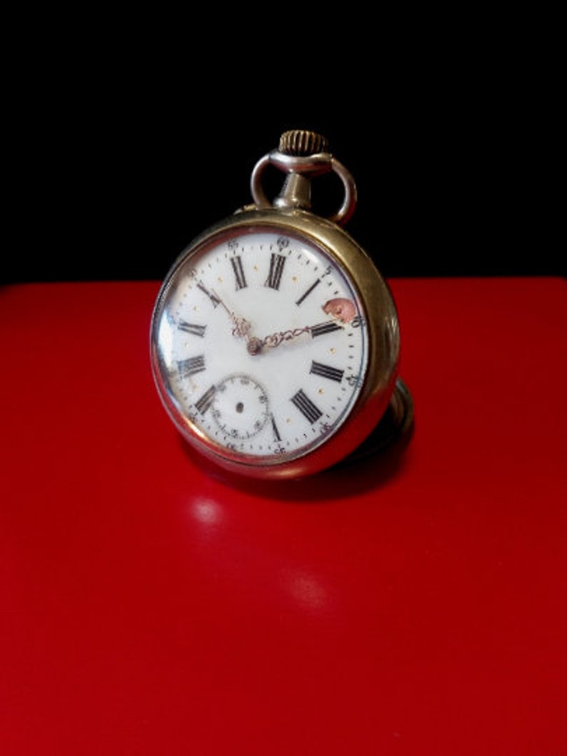 May include: A silver pocket watch with Roman numeral hour markers and a white face. The watch is on a red surface.
