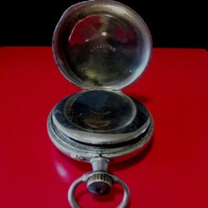 May include: A silver pocket watch with a hinged lid open, revealing the inner workings of the watch. The watch has a silver chain attached to the top.