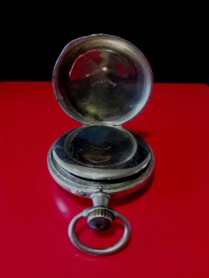 May include: A silver pocket watch with a hinged lid open, revealing the inner workings of the watch. The watch has a silver chain attached to the top.