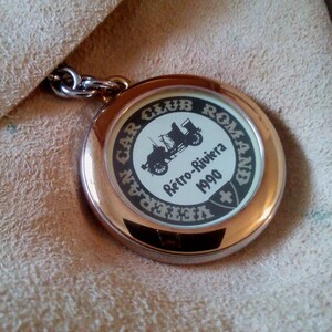 Peut inclure: Un porte-clés doré avec un charme rond bordé d'argent. Le charme présente une image en noir et blanc d'une voiture ancienne avec le texte "Veteran Car Club Romand Retro-Riviera 1990" et un drapeau suisse.