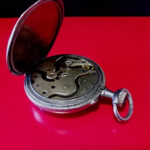 May include: A silver pocket watch with the back cover open, revealing the intricate clockwork mechanism. The watch is on a red surface.