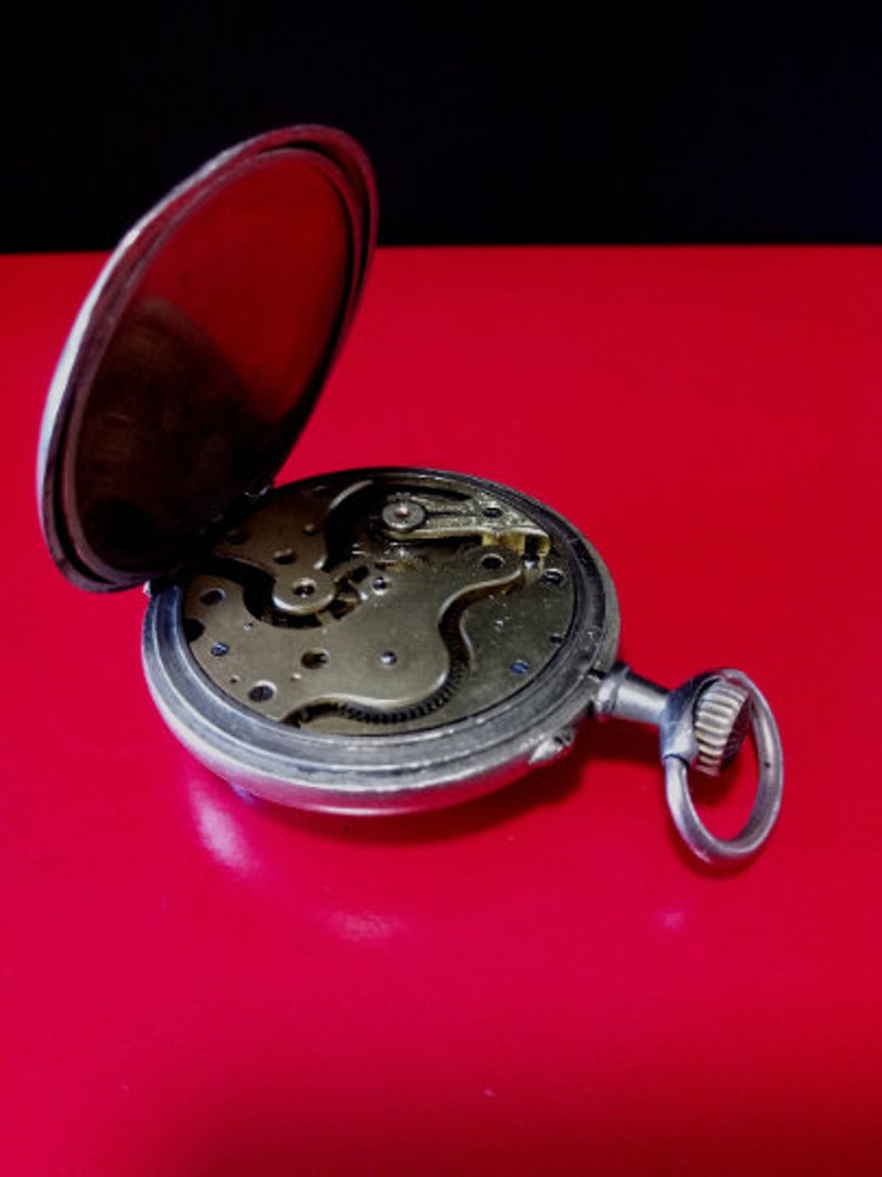May include: A silver pocket watch with the back cover open, revealing the intricate clockwork mechanism. The watch is on a red surface.