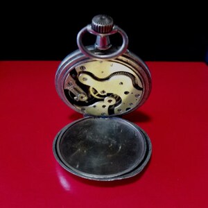 May include: Open silver pocket watch with a visible clockwork mechanism. The watch has a round, silver case and a hinged lid.