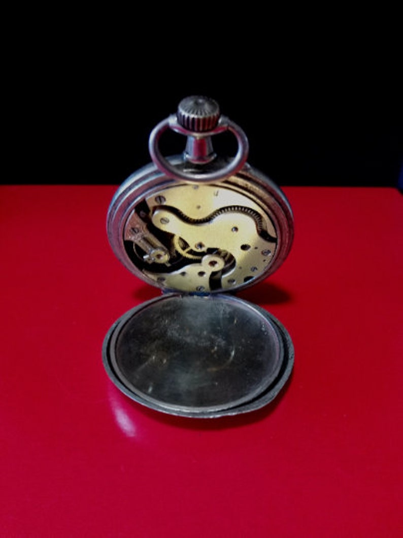 May include: Open silver pocket watch with a visible clockwork mechanism. The watch has a round, silver case and a hinged lid.