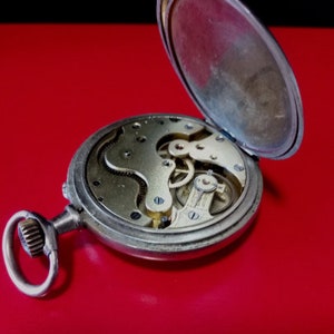 May include: A silver pocket watch with the back open, revealing the intricate clockwork mechanism. The watch has a simple design with a round face and a small, round knob on the side.