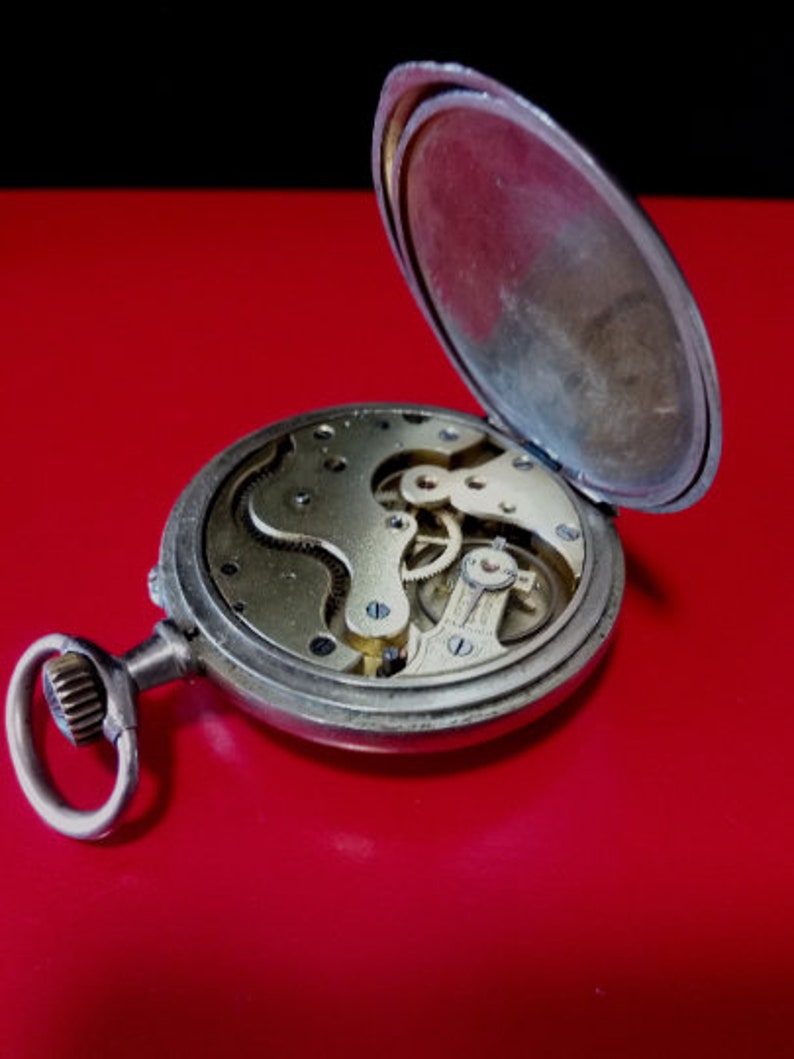 May include: A silver pocket watch with the back open, revealing the intricate clockwork mechanism. The watch has a simple design with a round face and a small, round knob on the side.
