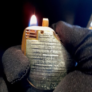 May include: A silver and gold lighter with a textured surface is being held in a gloved hand. The lighter is lit and has a flame coming out of the top.