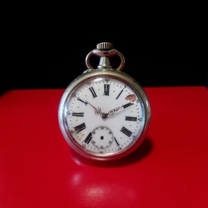 May include: A silver pocket watch with Roman numeral hour markers and a white face. The watch has a small second hand dial at the bottom of the face.
